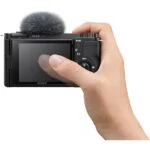 Hand holding a Sony mirrorless camera with a microphone attached, showcasing the rear screen.