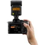 Person holding a Sony mirrorless camera with attached flash, displaying settings on the screen.