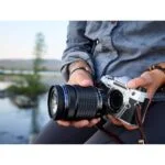 Person holding Olympus mirrorless camera with lens outdoors.