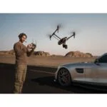 Man operating a DJI drone with remote control near a car in a desert landscape.