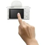 Person touching the screen of a white digital camera, showcasing camera features.