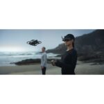 Person flying a drone on a beach using VR headset and controller.
