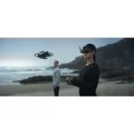 Person flying a drone on a beach using VR headset and controller.