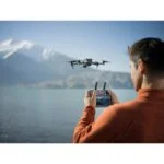 Man flying DJI drone over scenic lake with mountains in background.