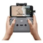 Hands holding DJI drone controller with smartphone display, aerial view of beach.