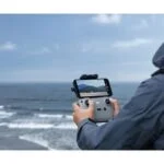 Person using a DJI drone controller by the sea, capturing aerial footage.