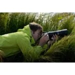 Photographer in green jacket using a DSLR camera with a telephoto lens in a grassy field.