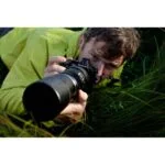 Photographer using Olympus camera with telephoto lens in grassy field.