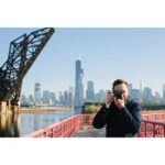 Man photographing city skyline with a digital camera on a bridge.
