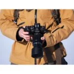 Person holding an Olympus camera with a zoom lens, wearing a brown jacket.