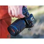 Person holding Olympus mirrorless camera with zoom lens outdoors.