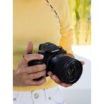 Person holding a Panasonic Lumix camera with a lens, outdoors.