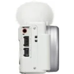 Side view of a white digital camera with microphone attachment, showing ports and controls.