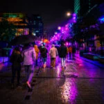 Night city scene with people walking under colorful lights and reflections on wet pavement.