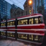 Red streetcar in urban setting with reflection, evening cityscape.