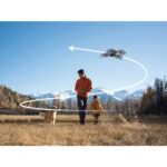 Man and child with dog in field, drone flying overhead, clear sky, mountains in background.