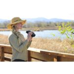 Woman using binoculars outdoors near a lake, wearing a straw hat.