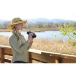 Woman using binoculars outdoors near a lake, wearing a straw hat.