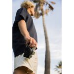 Person holding a mirrorless camera outdoors with a palm tree in the background.