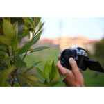 Person holding a Nikon mirrorless camera outdoors near green leaves.
