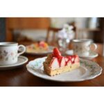 Slice of strawberry tart on a plate with teacups in the background.