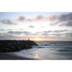 Sunset over ocean with rocky shore, people silhouetted against sky.