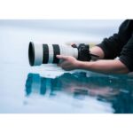 Person holding a professional camera with a large telephoto lens near water.