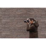 Photographer using a digital camera against a brick wall backdrop.