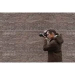 Photographer using a digital camera against a brick wall backdrop.