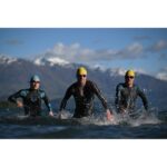 Three athletes in wetsuits and swim caps running through water with mountains in the background.